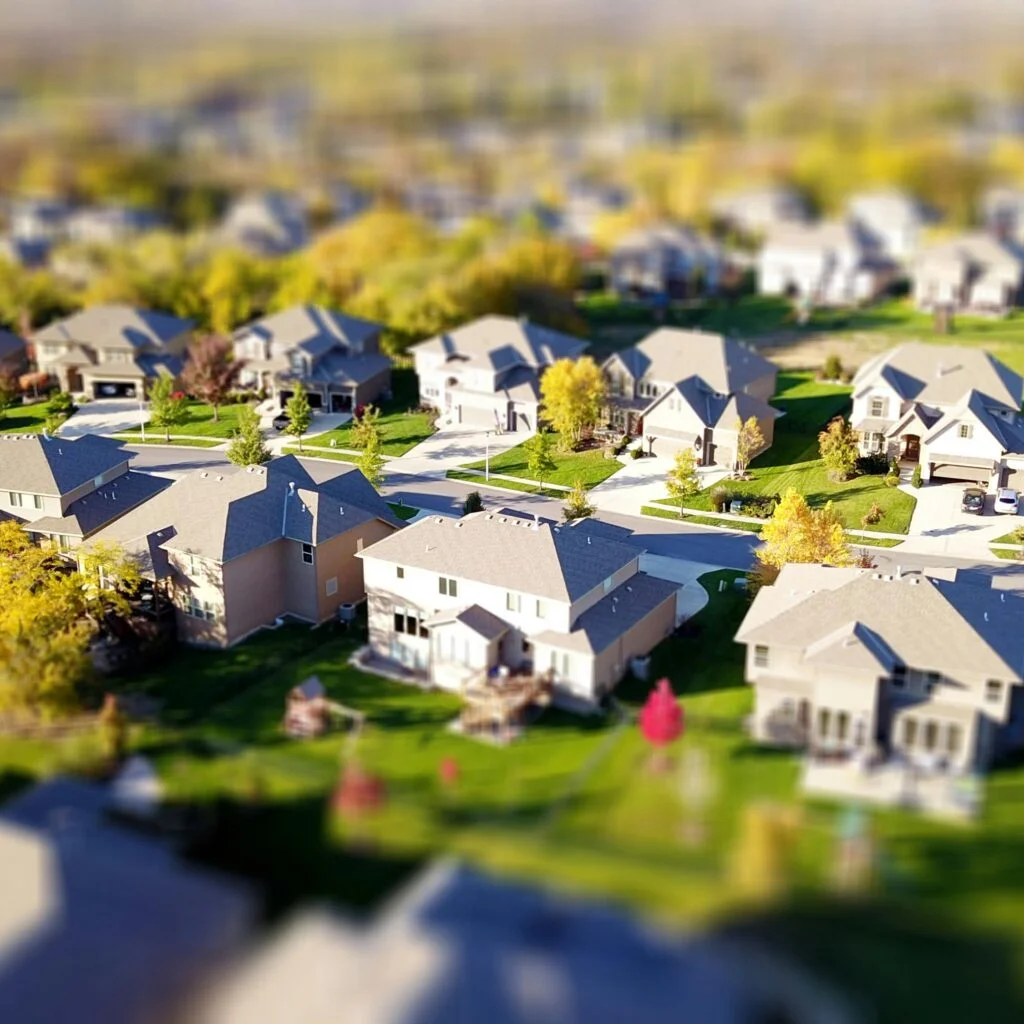 Tilt-shift aerial photo of a suburban neighborhood with green lawns and roads.